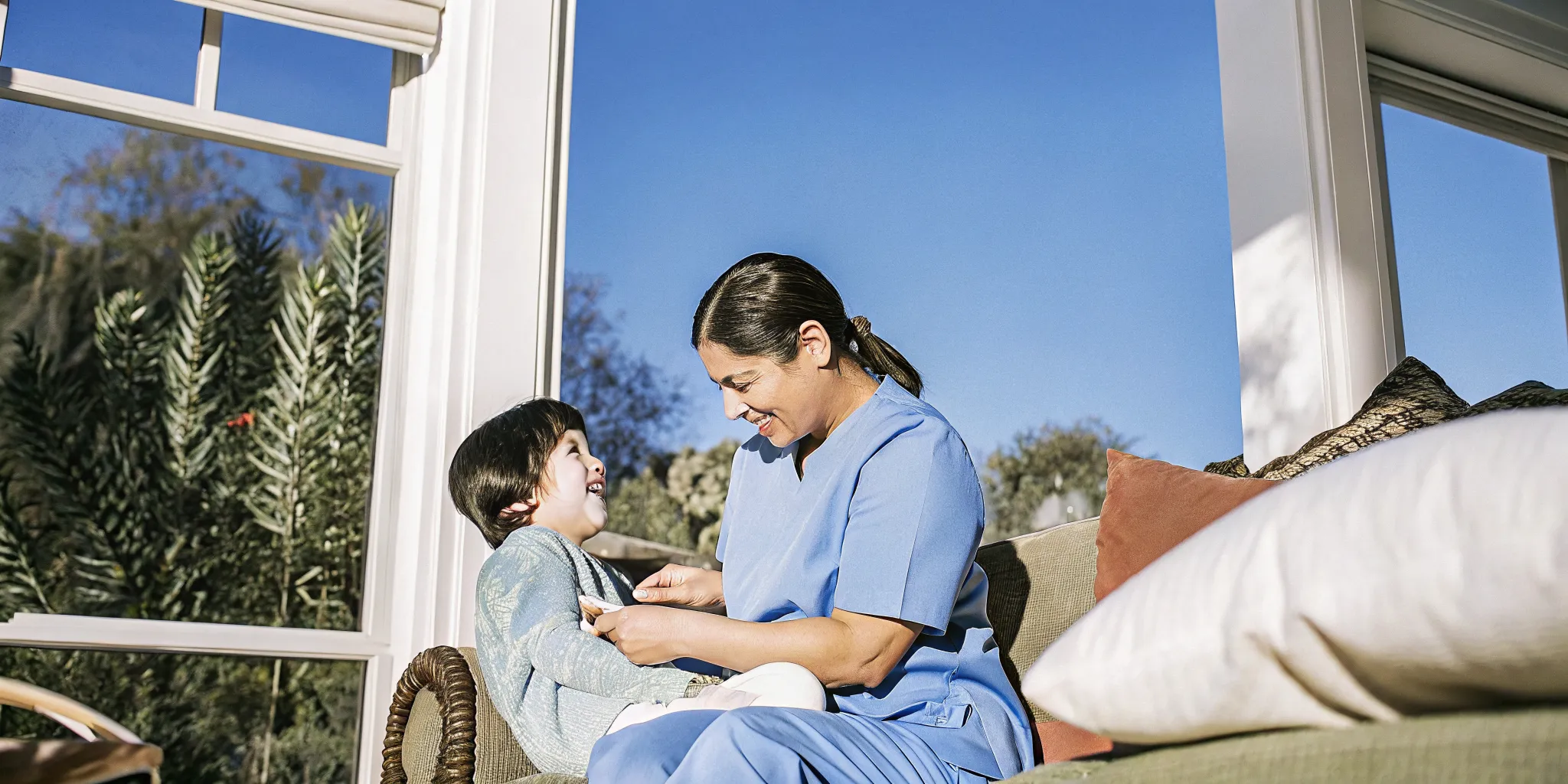 A pediatric home nurse gently assists a young child with Down syndrome in a warm, bright family home setting